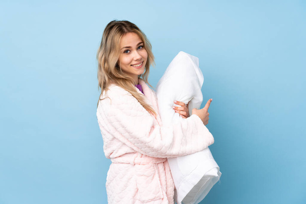 Teenager Russian girl in pajamas isolated on blue background pointing back - 写真・画像