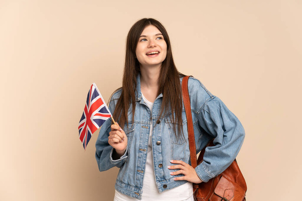 junges ukrainisches Mädchen mit einer Flagge des Vereinigten Königreichs isoliert auf beigem Hintergrund posiert mit den Armen an der Hüfte und lächelt - Foto, Bild