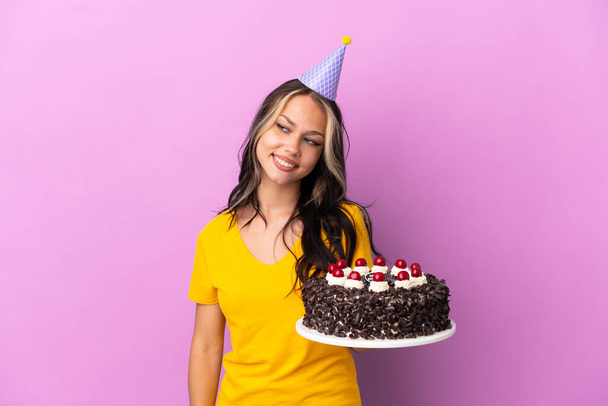 Adolescente russo menina segurando bolo de aniversário isolado no fundo roxo olhando para o lado e sorrindo - Foto, Imagem