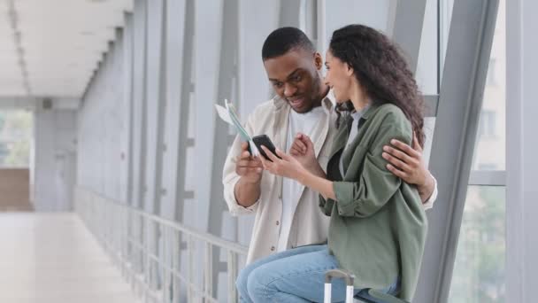 Multiracial couple african man and hispanic woman with plane tickets passports sitting in airport terminal using navigator app mobile phone watching photo from trip browsing network with smartphone - Footage, Video