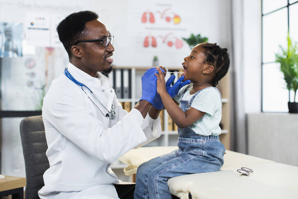 Black pediatrician examining throat of cute child at clinic - Foto, Imagen