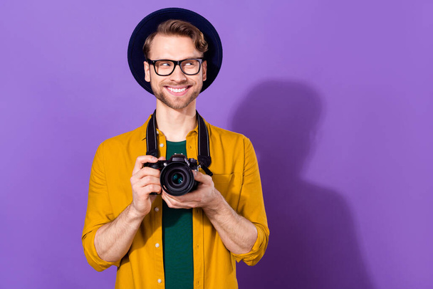 Portrait of nice cheerful curious guy holding in hands cam looking aside copy space isolated over bright violet purple color background - 写真・画像
