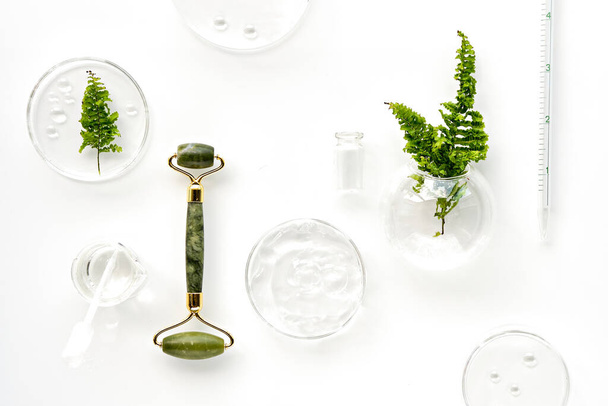 Self made moisturizer and green jade face roller. Exotic fern leaves and water drops on white background. Facial massage, handmade cosmetics. Monochromatic white green look. - Фото, зображення