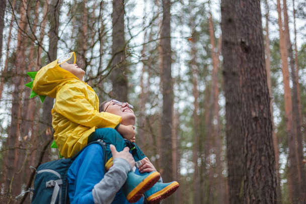 Mom and child walking in the forest after the rain in raincoats together, the child is sitting on the shoulders of mom, happily looking at the sky - Fotografie, Obrázek