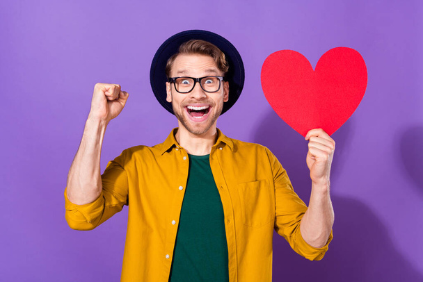 Photo of cheerful charming victorious young man winner raise fists hold red paper heart isolated on violet color background - Fotografie, Obrázek