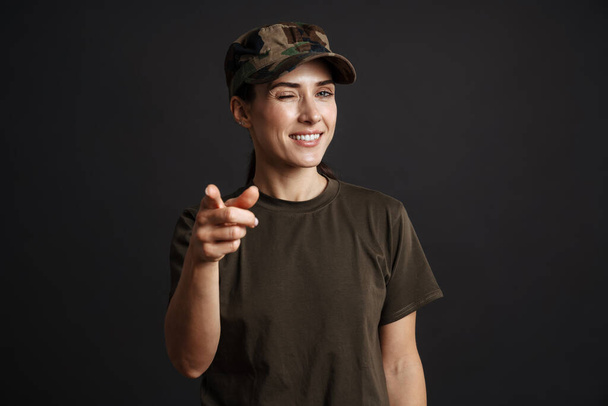 Happy soldier woman winking and pointing finger at camera isolated over black background - Foto, afbeelding
