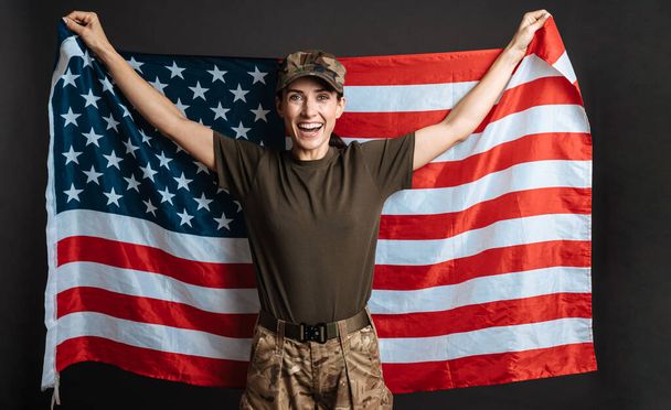 Delighted soldier woman smiling while posing with american flag isolated over black background - Fotoğraf, Görsel
