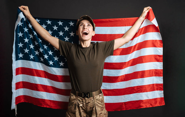 Delighted soldier woman smiling while posing with american flag isolated over black background - Zdjęcie, obraz