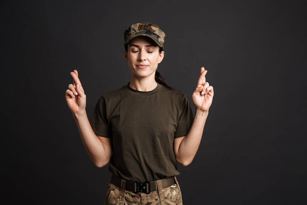 Calm soldier woman holding fingers crossed for good luck isolated over black background - Фото, зображення