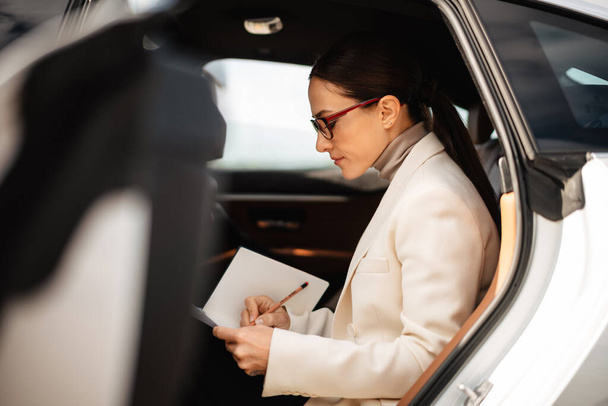 Beautiful focused businesswoman writing down notes while sitting in car - Valokuva, kuva