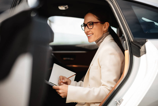 Beautiful happy businesswoman smiling and writing down notes while sitting in car - Foto, Imagem