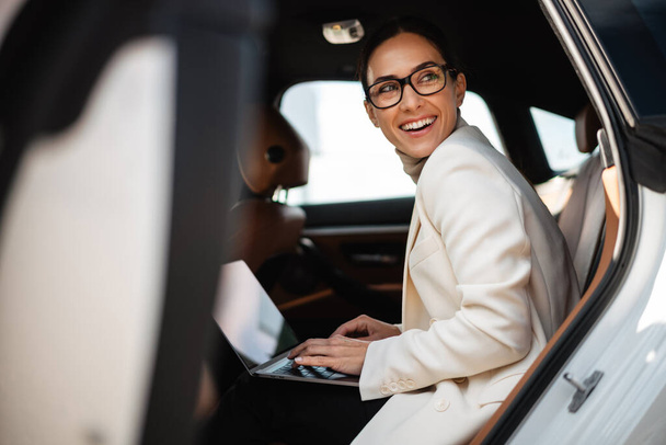 Beautiful smiling businesswoman working with laptop while sitting in car - Foto, Imagen