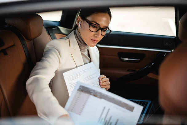 Όμορφη εστιασμένη επιχειρηματίας σε ακουστικά που εργάζονται με φορητό υπολογιστή και χαρτιά, ενώ κάθεται στο αυτοκίνητο - Φωτογραφία, εικόνα