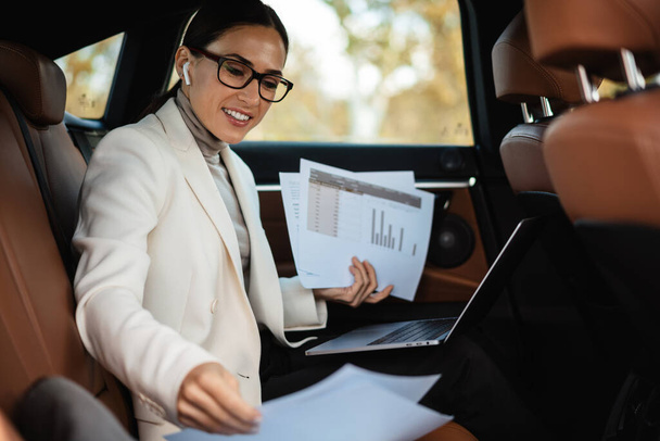 Beautiful smiling businesswoman in earphones working with laptop and papers while sitting in car - Photo, image