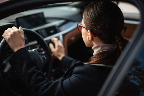 Beautiful focused businesswoman in earphones and eyeglasses driving car - Фото, зображення