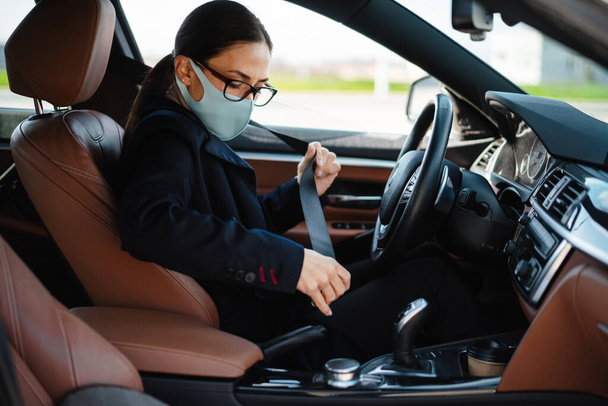 Image of a pretty business woman sitting in a car in face mask and fastening her seabelt - Foto, Imagen
