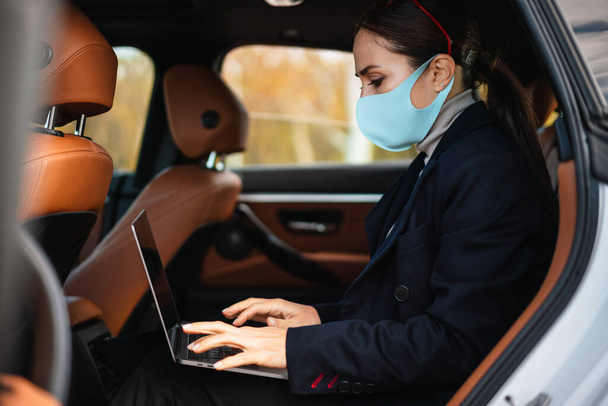 Beautiful confident focused businesswoman wearing face mask working with laptop in car - 写真・画像
