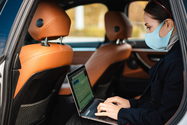 Beautiful confident focused businesswoman wearing face mask working with laptop in car - Zdjęcie, obraz
