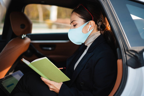 Image of beautiful business woman sitting in a car in face mask while holding notebook - Фото, зображення