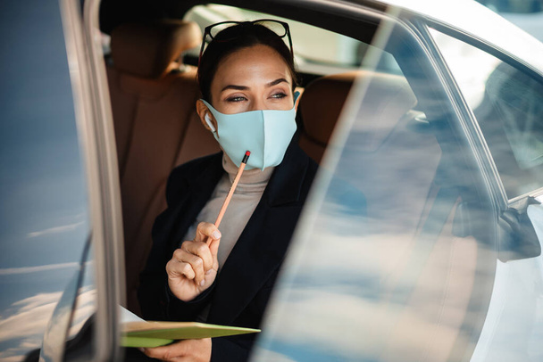 Photo of concentrated business woman sitting in a car in face mask while holding notebook - Φωτογραφία, εικόνα