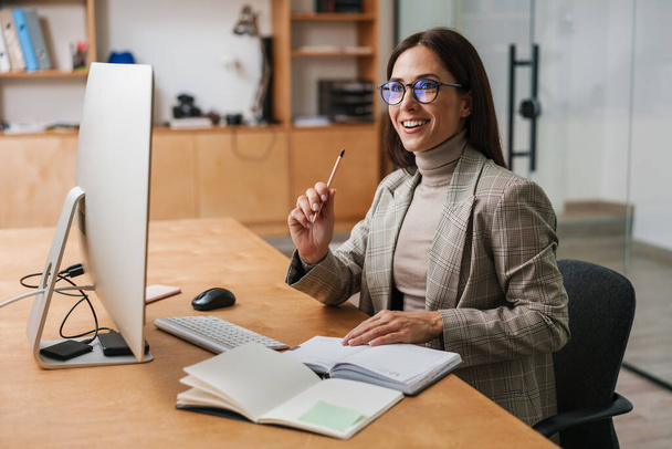 Joyful charming woman in eyeglasses working with computer and planner while sitting in office - Фото, зображення