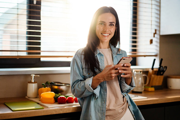 Smiling attractive young woman texting on mobile phone in the kitchen - Fotó, kép
