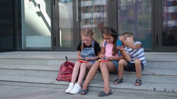  Drei Kinder mit Rucksack sitzen und spielen Popit und Handy - Filmmaterial, Video