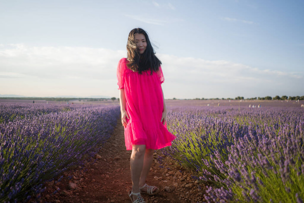 young Asian woman outdoors at lavender flowers field - happy and beautiful Korean girl in sweet Summer magenta dress enjoying holidays relaxed on purple floral meadow - 写真・画像