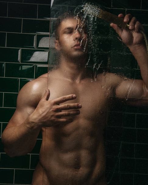 Naked muscular young guy pouring water from shower on glass in bathroom - Fotó, kép