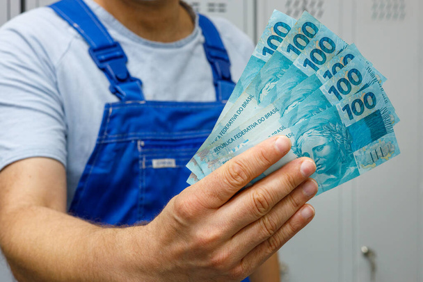 brasilianischer Arbeiter zeigt Geld. Banknotenbündel zu hundert Reais, Gehälter von Fachkräften und Arbeitern - Foto, Bild