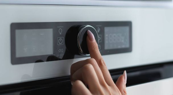 Γυναικείο δάχτυλο αγγίζει το κουμπί του σύγχρονου πίνακα του ηλεκτρικού φούρνου. Τεχνολογική έννοια. Εστίαση στο γυναικείο δάχτυλο. - Φωτογραφία, εικόνα