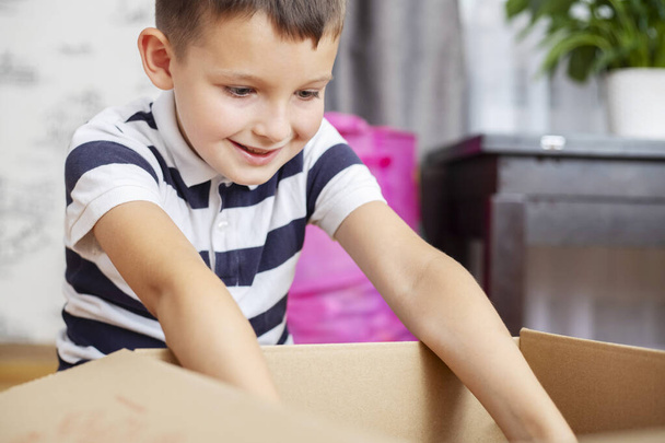 Ένα αγόρι ξεπακετάρει χαρτόκουτο (κουτί δώρου ή δέμα) στο σπίτι. Παράδοση και υπηρεσία έννοια. Online αγορές. - Φωτογραφία, εικόνα