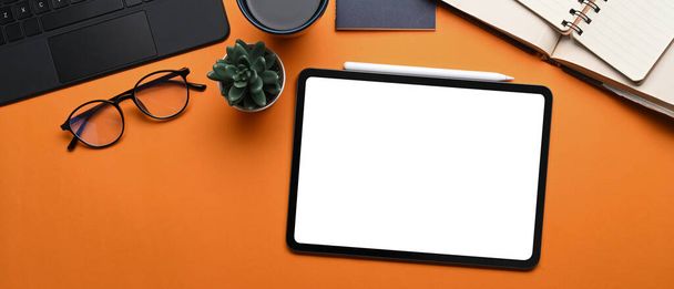 Vista superior de maqueta de tableta digital con pantalla de lank, lápiz lápiz óptico, gafas, cuaderno y planta sobre fondo naranja. - Foto, imagen