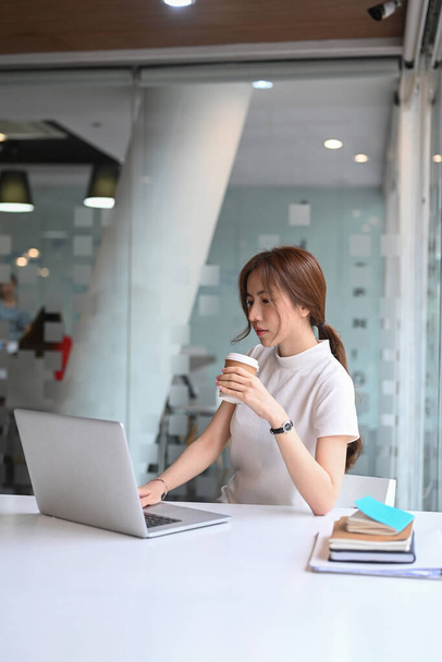Πορτρέτο της ευχάριστης επιχειρηματία κρατώντας φλιτζάνι καφέ και εργάζονται σε φορητό υπολογιστή. - Φωτογραφία, εικόνα