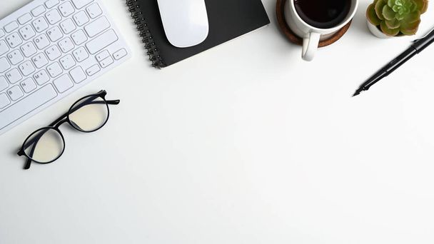 Pohled shora na bílý kancelářský stůl s šálkem kávy, brýlemi, klávesnicí, notebookem a kopírovacím prostorem. - Fotografie, Obrázek