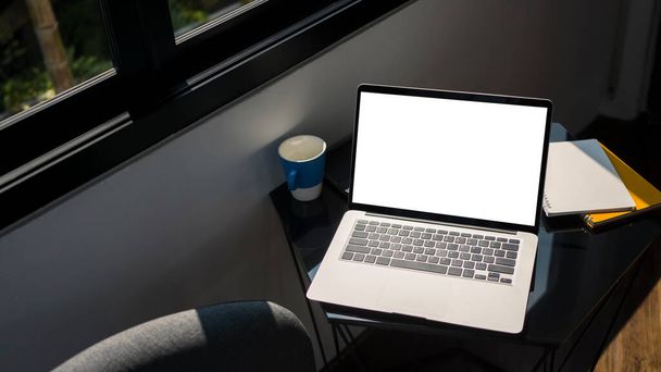 Computadora portátil simulada con pantalla en blanco, papeleo y taza de café en mesa negra en la oficina. - Foto, Imagen