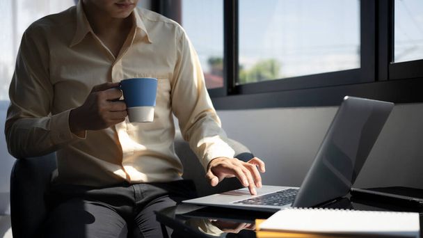 自信のあるビジネスマンのコーヒーカップを保持し、ラップトップコンピュータ上で作業の作物のショット. - 写真・画像