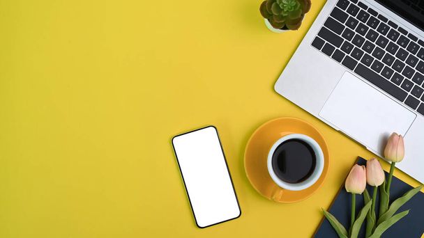 Horní pohled na ženský pracovní prostor s notebookem, šálkem kávy, květinami, chytrým telefonem a kopírovacím prostorem na žlutém pozadí. - Fotografie, Obrázek