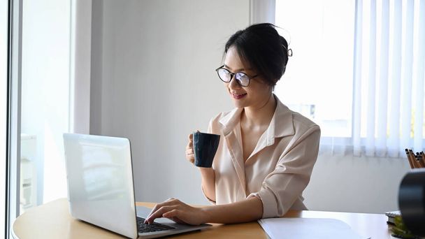 Smiling businesswoman in eyeglasses holding coffee cup and checking email on laptop computer. - Foto, Imagem