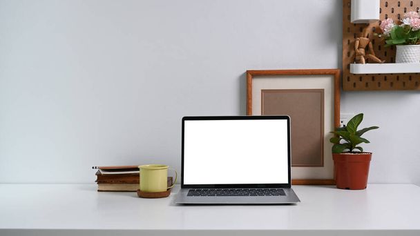 Jednoduché pracoviště s notebookem, knihami, šálkem kávy, domem a prázdným fotografickým rámečkem na bílém stole. - Fotografie, Obrázek