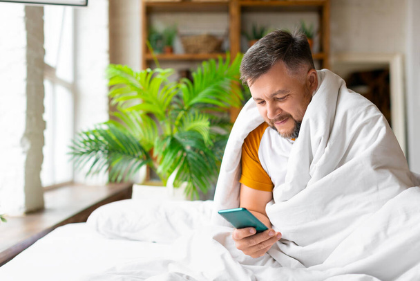 Középkorú férfi az ágyban ellenőrzi az üzeneteit okostelefonon ébredés után. A közösségi hálózatok koncepciójának függősége. - Fotó, kép