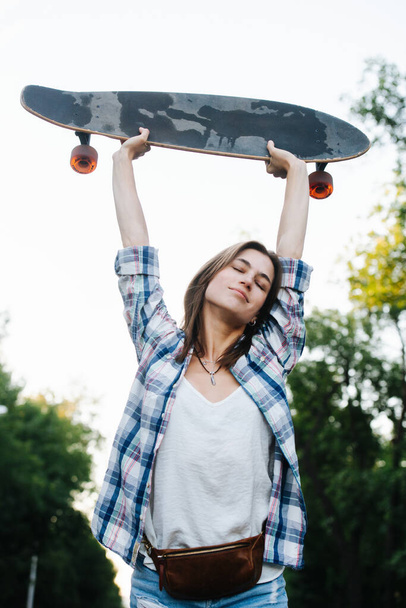 Tired happy woman stretching with a skateboard in hands. Low angle. She's wearing open checkered shirt atop regular white one. Eyes closed. - Zdjęcie, obraz