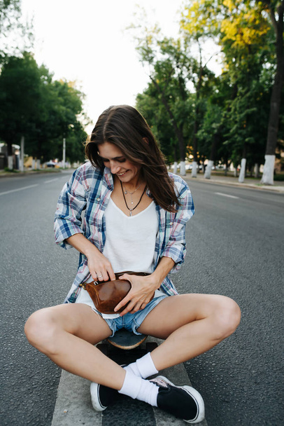 Carefree woman sitting cross legged in the middle of an empty city road, looking for something in her brown leather fanny pack. She's wearing checkered shirt and mini jeans shorts. - Zdjęcie, obraz