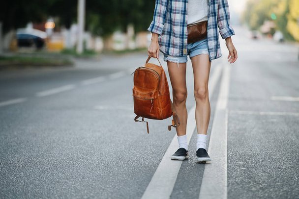 Lower part of a woman walking on the road, holding brown leather backpack. No head. She's wearing checkered shirt and mini jeans shorts. - Foto, afbeelding