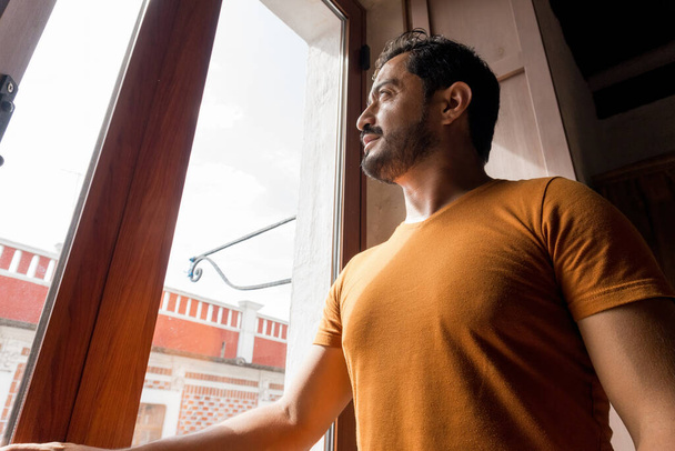 Egy jóképű, szakállas spanyol férfi narancssárga pólóban, kinézett az ablakon. - Fotó, kép