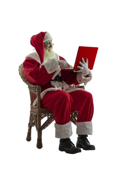 Santa Claus on white background isolated. Senior male actor old man with a real white beard in the role of Father Christmas sitting in a wicker willow chair with red laptop. - Photo, Image