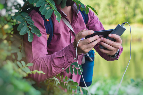 Tourist tart egy okostelefon hordozható töltő a kezében. Egy Power Bank-os férfi a telefont a természet és az erdő hátterébe helyezi. - Fotó, kép