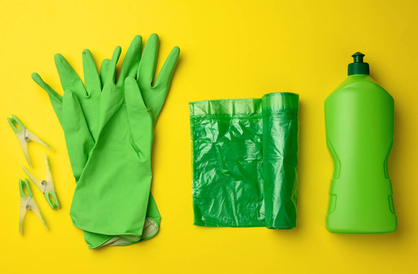 резиновые зеленые перчатки для чистки, мусорный бак пластиковый рулон мешок и пластиковая бутылка с моющим средством на желтом фоне, набор - Фото, изображение