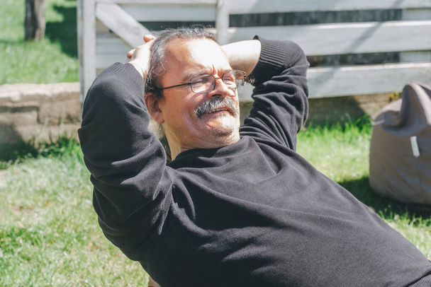 Портрет седого взрослого мужчины в очках и усах, сидящего на бобовом мешке и улыбающегося с руками за головой - Фото, изображение