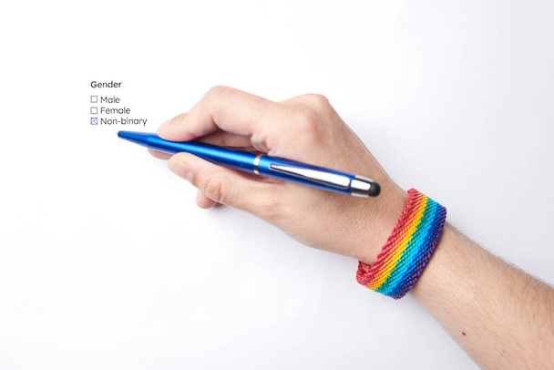 Hand einer unkenntlichen Person, die die nicht binäre Option auf einem Formular markiert, das diese Option der Geschlechtsidentität enthält, trägt ein Armband mit der Regenbogenfahne, Symbol der lgbt-Gemeinschaft. - Foto, Bild